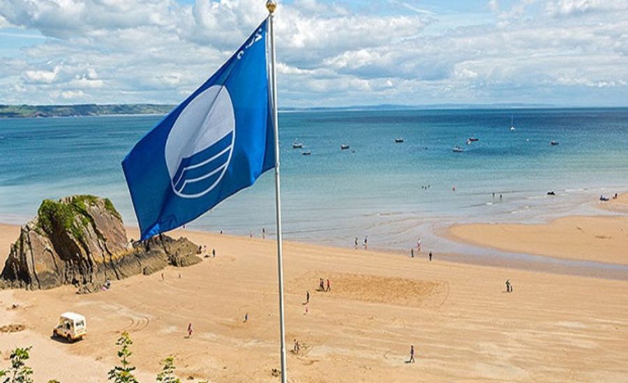A blue flag with a white wave symbol flies above a sandy beach with scattered people, rocky formations, and calm blue sea under a partly cloudy sky.