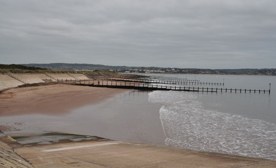 A cloudy day at a sandy beach with gentle waves, groynes stretching into the sea, and a distant coastline visible under an overcast sky.