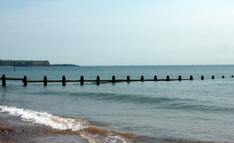 A calm sea with gentle waves near the shore. Wooden groynes extend out from the beach into the water. The sky is mostly clear, and distant land is visible on the horizon.