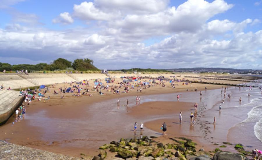 A sandy beach with many people relaxing, playing, and walking along the shoreline. Some are near the water, while others are sitting on the sand. The sky is partly cloudy with some greenery and concrete steps in the background.