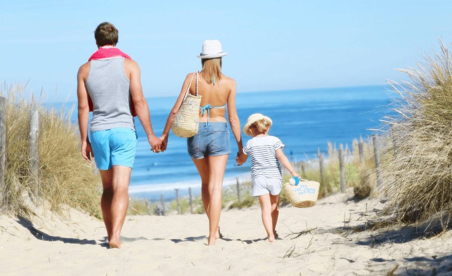 A family of three, wearing summer clothes and hats, walks hand in hand down a sandy path toward the beach and ocean on a sunny day.